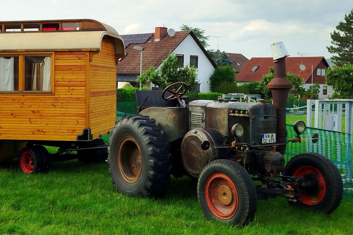 Ein Lanz D9506 in Patina mit einem Holzanhänger steht auf einer Wiese.